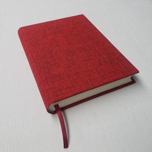 May include: A red hardcover journal with a textured cover and a matching ribbon bookmark. The journal's pages are cream-colored, and the cover has a subtle, dark flecked pattern. The book is closed and resting on a white surface.