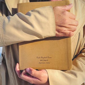 May include: A brown leather-bound journal with the inscription "Paula Elizabeth Torres SKE Class 3 Matthew-Acts" on the front cover.