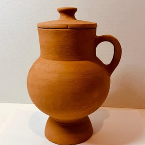 May include: A terracotta pitcher with a rounded body, a wide neck, and a matching lid featuring a small knob handle. The vessel has a single curved handle on the side and sits on a flared pedestal base, showcasing a matte, earthy orange finish.