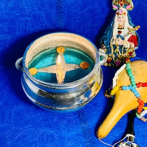 May include: A silver-toned metal pot with two handles containing a teal liquid and a floating star-shaped wick holder with a lit flame. Beside it sits a small figurine of a person in blue and red robes and a tan gourd instrument with colorful beads.