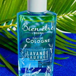 Puede incluir: Una botella de vidrio transparente de "Bien-être Eau de Cologne" con tapón blanco. La etiqueta presenta texto en francés, incluyendo "Lavande Sauvage" e ilustraciones florales. La botella contiene un líquido azul claro, sobre fondo azul.