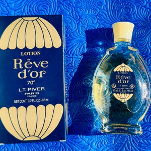 May include: A vintage bottle of "Rêve d'or" lotion and its matching box. The bottle is clear with a yellow cap, and the box is navy blue with gold accents. The text "Rêve d'or" is visible on both the bottle and the box. The bottle contains 97 ml.