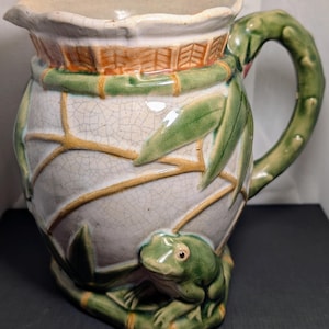 May include: A ceramic pitcher with a cream-colored body, adorned with green bamboo and leaves, and a frog at the base. The handle is green, and the rim features a woven design. The pitcher has a crackle glaze finish.