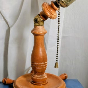 May include: A wooden desk lamp with a nautical theme. The lamp features a base shaped like a ship's wheel, a central column, and an adjustable top with a brass light socket. The lamp has a pull chain with a small bell.
