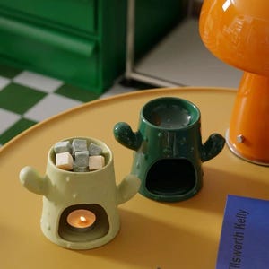 Puede incluir: Dos calentadores de cera derretida de cerámica con forma de cactus. Uno es verde y el otro es verde claro. Ambos tienen un pequeño agujero en el centro para una vela de té. El calentador verde claro tiene cera derretida dentro y una vela de té encendida.