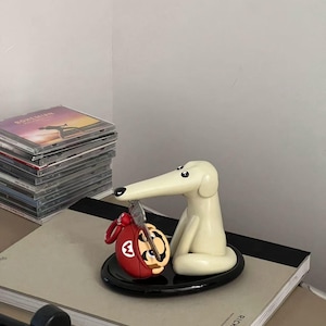 May include: A white ceramic dog figurine with a long snout holding a red and black Mario character keychain. The figurine is sitting on a black circular base and is on top of a gray book with the text "RICK OWENS" printed on the cover.