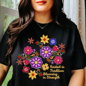 May include: Black t-shirt featuring a floral design with purple, pink, yellow, and blue flowers. The text "Rooted in Tradition Blooming in Strength" is printed in gold. The shirt is short-sleeved.