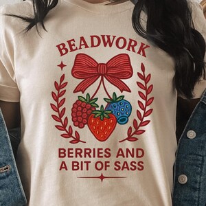 Peut inclure: T-shirt crème clair avec l'inscription "BEADWORK BERRIES AND A BIT OF SASS". Le motif comprend un nœud rouge, des baies et une couronne de laurier. La chemise est portée avec une veste en jean et un pantalon blanc.