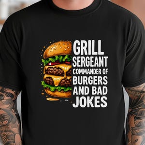 May include: Black t-shirt with a graphic of a double-decker burger and the text "GRILL SERGEANT COMMANDER OF BURGERS AND BAD JOKES" in white. The burger has two patties, lettuce, and cheese. The shirt is a crew neck.