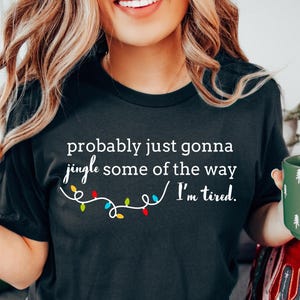 K&ouml;nnte beinhalten: Schwarzes T-Shirt mit dem Text "probably just gonna jingle some of the way I'm tired." Die Grafik zeigt eine Lichterkette mit bunten Weihnachtslichtern. Das Model tr&auml;gt hellblaue Jeansshorts.