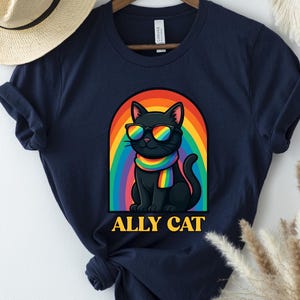 Peut inclure: T-shirt bleu marine avec un chat noir portant des lunettes de soleil et une écharpe arc-en-ciel, sur fond d'arc-en-ciel. Le texte "ALLY CAT" est en dessous du chat. Un chapeau de paille et une décoration murale sont en arrière-plan.