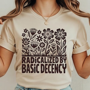 Peut inclure: T-shirt beige avec un motif floral marron et le texte "RADICALIZED BY BASIC DECENCY". Le t-shirt est en matière douce et convient aux tenues décontractées. Le modèle porte un jean bleu clair.