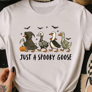 K&ouml;nnte beinhalten: Wei&szlig;es T-Shirt mit einem Halloween-Design. Die Grafik zeigt vier Cartoon-G&auml;nse in gruseligen Kost&uuml;men, darunter der Sensenmann und ein Skelett. Der Text "JUST A SPOOKY GOOSE" ist unter den G&auml;nsen aufgedruckt.