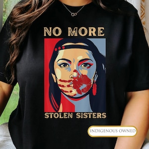 May include: Black t-shirt with a graphic design featuring a woman's face with a red handprint over her mouth. The text "NO MORE" is above the image, and "STOLEN SISTERS" is below. The shirt also has the text "INDIGENOUS OWNED".