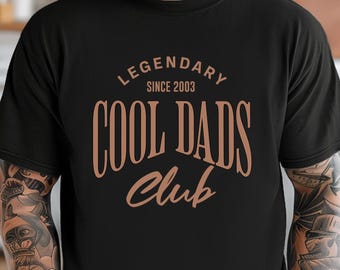 May include: Black t-shirt with the text "LEGENDARY SINCE 2003 COOL DADS Club" in a copper-colored font. The shirt has a crew neck and short sleeves. The image shows the shirt being worn.