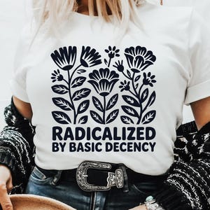 Peut inclure: T-shirt blanc avec un motif floral bleu marine et le texte "RADICALIZED BY BASIC DECENCY". Le t-shirt est porté avec un jean bleu, un cardigan noir et blanc et un chapeau marron.