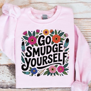 May include: Pale pink sweatshirt featuring the phrase "GO SMUDGE YOURSELF" in black, framed by vibrant floral and leaf designs. The sweatshirt has a crew neck.