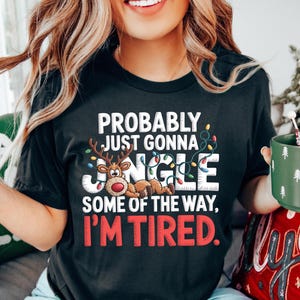 May include: Black t-shirt with the text "PROBABLY JUST GONNA JINGLE SOME OF THE WAY, I'M TIRED." The design includes a reindeer and Christmas lights. The person is holding a green mug with white tree designs.