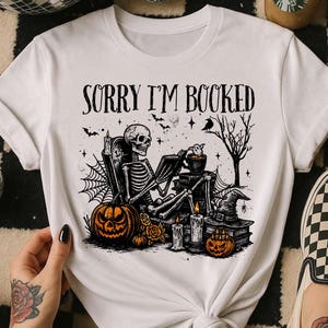 Peut inclure: T-shirt blanc avec un motif sur le thème d'Halloween. Le motif représente un squelette, des citrouilles, des bougies et le texte "SORRY I'M BOOKED". Le t-shirt est porté avec des chaussures à enfiler à carreaux.