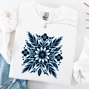 Peut inclure: Sweat-shirt blanc orné d'un motif floral symétrique noir et bleu. Le motif comprend des feuilles, des fleurs et des points stylisés, créant un motif géométrique. Un collier argenté est visible.