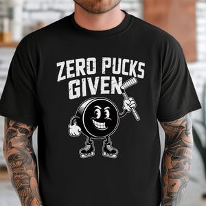 May include: Black t-shirt with the text "ZERO PUCKS GIVEN" in white. A cartoon hockey puck character with a smiling face and hockey stick is also featured. The shirt is a crew neck.