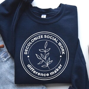 May include: Navy blue sweatshirt with a white circle graphic that says "Decolonize Social Work Difference Maker" and a small branch design inside the circle.