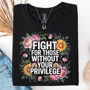 May include: Black t-shirt featuring the text "FIGHT FOR THOSE WITHOUT YOUR PRIVILEGE" within a floral wreath of pink peonies, yellow sunflowers, and green leaves. A rainbow graphic is below the text. A silver necklace with a sun pendant is visible.