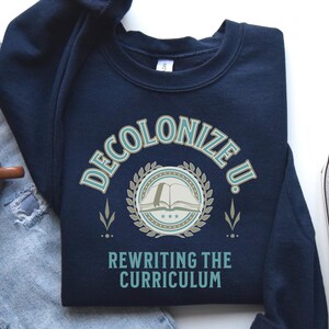 May include: Navy blue sweatshirt with the text "Decolonize U. Rewriting the Curriculum" in white and teal lettering. The sweatshirt features a teal and white graphic of an open book with stars and a laurel wreath. The text "Indigenous Owned" is in white lettering on a black background in the bottom right corner.