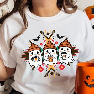 May include: White t-shirt featuring a Halloween-themed graphic. The design includes three cartoon ghosts wearing orange beanies and holding coffee cups. The shirt also has a geometric pattern and bat silhouettes. An orange mug with a jack-o'-lantern face is also visible.