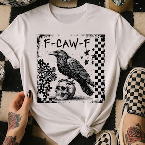 May include: White t-shirt with a black raven design, text "F-CAW-F", perched on a skull. The design includes stars, flowers, and a checkered pattern. The shirt is styled with checkered slip-on shoes.