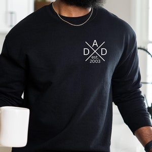 Peut inclure: Sweat-shirt à manches longues noir avec un motif blanc en forme de croix avec les lettres "A" et "D" de chaque côté et le texte "est. 2003" en dessous.