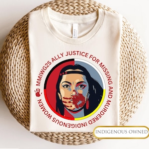 May include: A beige t-shirt with a graphic of a woman's face with a red handprint over her mouth. The graphic is surrounded by a circle with the text "MMIW2S ALLY JUSTICE FOR MISSING AND MURDERED INDIGENOUS WOMEN" in black text. The text "INDIGENOUS OWNED" is in black text at the bottom of the shirt.