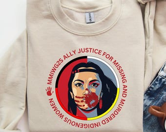 May include: Sand-colored sweatshirt with a circular graphic featuring a woman's face, half red and half black, with a handprint over her mouth. Text around the graphic reads "MMIWG2S ALLY JUSTICE FOR MISSING AND MURDERED INDIGENOUS WOMEN". The text "INDIGENOUS OWNED" is also visible.
