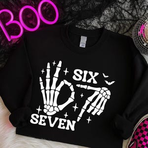 May include: Black sweatshirt with a white skeleton hand design. The design includes the words "SIX" and "SEVEN", along with star and bat graphics. The sweatshirt is a Halloween-themed apparel item.