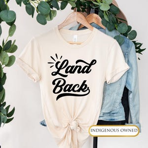 May include: A beige t-shirt with the text "Land Back" in black stylized lettering. The shirt is tied in a knot at the bottom and has a denim jacket draped over it. The text "Indigenous Owned" is printed in gold on a black rectangle at the bottom of the image.