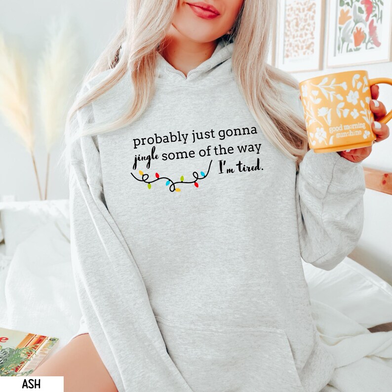 K&ouml;nnte beinhalten: Ein hellgrauer Kapuzenpullover mit dem Text "probably just gonna jingle some of the way I'm tired." Der Text ist schwarz, mit einer Lichterkette. Der Kapuzenpullover hat eine Vordertasche und eine Kapuze.