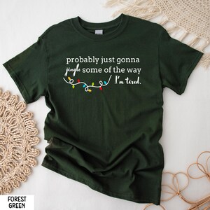 K&ouml;nnte beinhalten: Forstgr&uuml;nes T-Shirt mit dem Text "probably just gonna jingle some of the way / I'm tired." Unter dem Text befindet sich eine Lichterkette mit bunten Weihnachtslichtern. Das Shirt ist einfarbig.