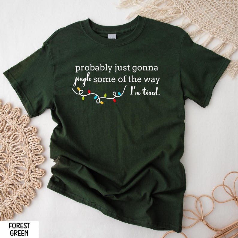 K&ouml;nnte beinhalten: Forstgr&uuml;nes T-Shirt mit dem Text "probably just gonna jingle some of the way / I'm tired." Unter dem Text befindet sich eine Lichterkette mit bunten Weihnachtslichtern. Das Shirt ist einfarbig.