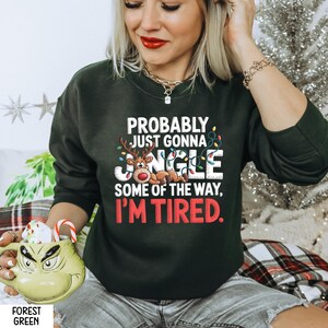 May include: Forest green sweatshirt with the text "PROBABLY JUST GONNA JANGLE SOME OF THE WAY, I'M TIRED." The design includes a reindeer, Christmas lights, and the word "JANGLE" in a festive font. A mug with a Grinch face is in the foreground.