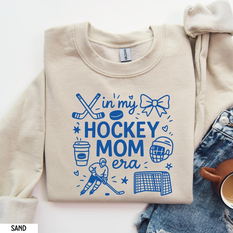 May include: Sand-colored sweatshirt with the phrase "In my Hockey Mom Era" in blue. The design includes hockey sticks, a puck, a bow, a coffee cup, a hockey player, a helmet, a net, and stars. The sweatshirt is made of a soft material.
