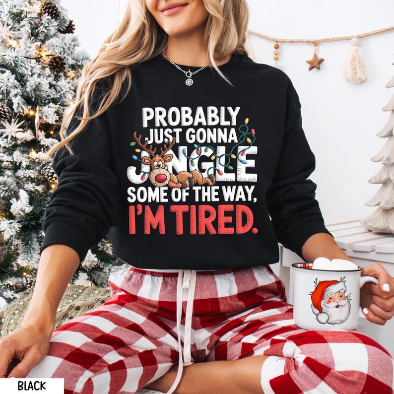 May include: Black sweatshirt with the text "PROBABLY JUST GONNA JINGLE SOME OF THE WAY, I'M TIRED." The graphic includes a reindeer and Christmas lights. The person is wearing red and white plaid pajama bottoms and holding a mug with a Santa Claus design.