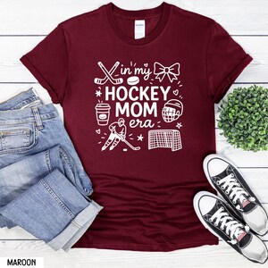 May include: Maroon t-shirt with the text "In my Hockey Mom Era" in white. The design includes hockey sticks, a puck, a bow, a coffee cup, a hockey player, a helmet, and a net. The shirt is paired with blue jeans and black and white sneakers.