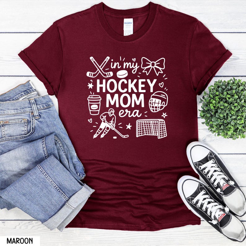 May include: Maroon t-shirt with the text "In my Hockey Mom Era" in white. The design includes hockey sticks, a puck, a bow, a coffee cup, a hockey player, a helmet, and a net. The shirt is paired with blue jeans and black and white sneakers.