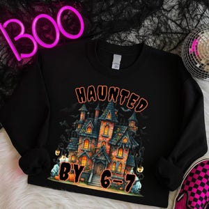 Peut inclure: Sweat-shirt noir avec un motif d'Halloween. Le motif représente une maison hantée avec le texte "HAUNTED BY 6-7". Le mot "BOO" est en néon rose en arrière-plan. Une boule disco et une paire de chaussures à carreaux roses et noirs sont également en arrière-plan.