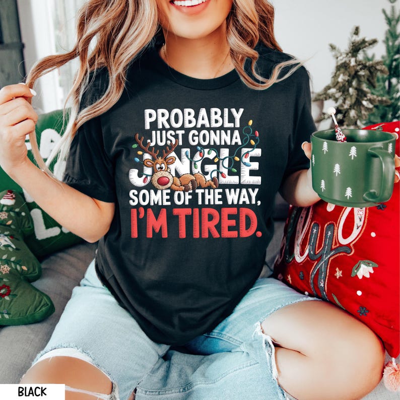 May include: Black t-shirt with the text "PROBABLY JUST GONNA JINGLE SOME OF THE WAY, I'M TIRED." The design includes a reindeer and Christmas lights. The person is holding a green mug with white tree designs.