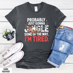 May include: Dark grey t-shirt with the text "PROBABLY JUST GONNA JINGLE SOME OF THE WAY, I'M TIRED." A reindeer graphic is featured in the design. White trainers and blue jeans are also visible.