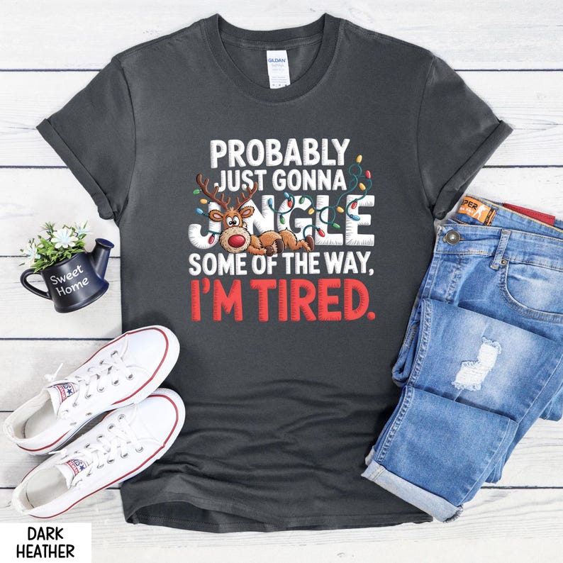 May include: Dark grey t-shirt with the text "PROBABLY JUST GONNA JINGLE SOME OF THE WAY, I'M TIRED." A reindeer graphic is featured in the design. White trainers and blue jeans are also visible.