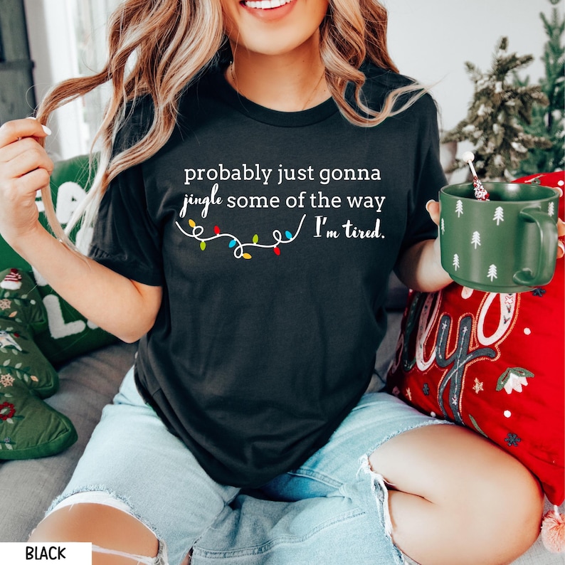 K&ouml;nnte beinhalten: Schwarzes T-Shirt mit dem Text "probably just gonna jingle some of the way I'm tired." Die Grafik zeigt eine Lichterkette mit bunten Weihnachtslichtern. Das Model tr&auml;gt hellblaue Jeansshorts.