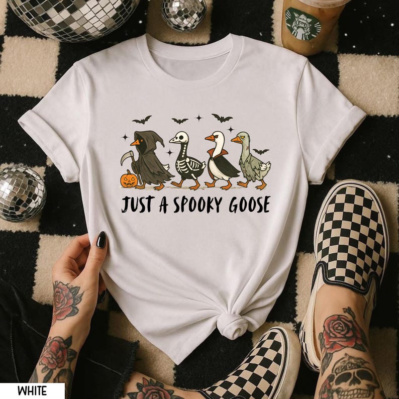 K&ouml;nnte beinhalten: Wei&szlig;es T-Shirt mit einem Halloween-Design. Die Grafik zeigt vier Cartoon-G&auml;nse in gruseligen Kost&uuml;men, darunter der Sensenmann und ein Skelett. Der Text "JUST A SPOOKY GOOSE" ist unter den G&auml;nsen aufgedruckt.