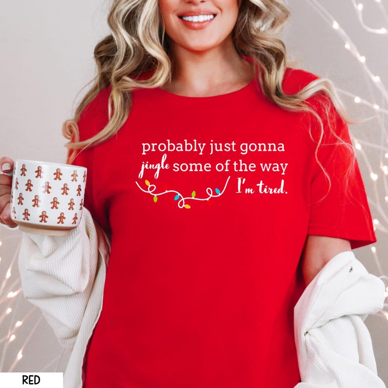 K&ouml;nnte beinhalten: Ein rotes T-Shirt mit dem Text "probably just gonna jingle some of the way I'm tired." Das Shirt zeigt eine Lichterkette mit bunten Weihnachtslichtern. Eine wei&szlig;e Tasse mit Lebkuchenm&auml;nnern wird im Bild gehalten.
